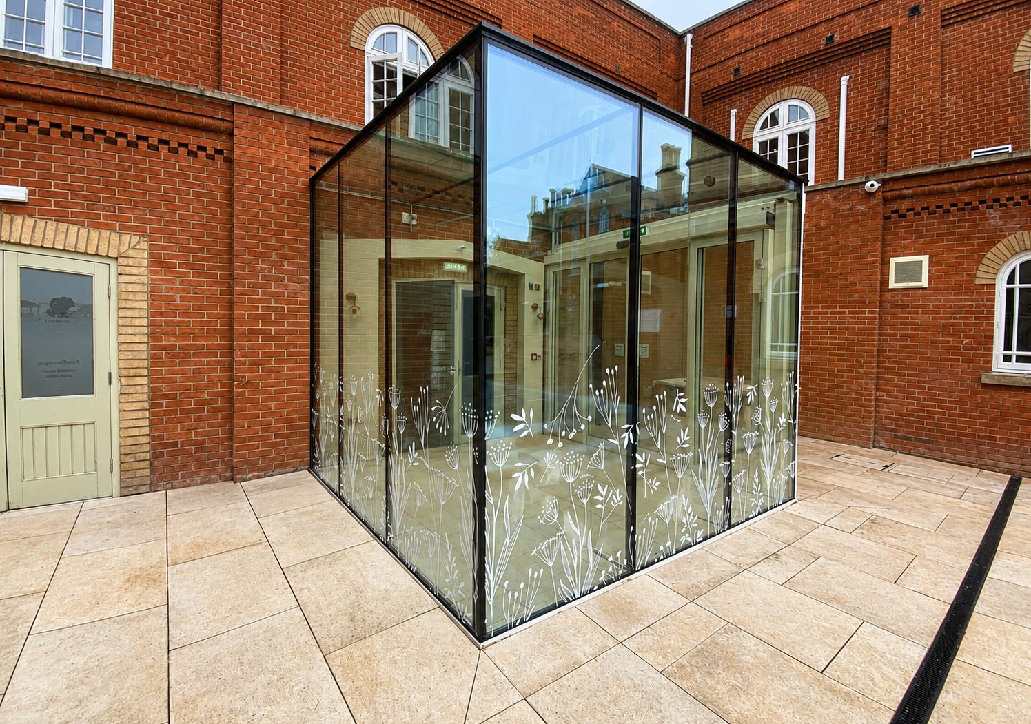 Glass-walled atrium extension with white floral etching along the bottom, set in a red-brick courtyard with beige tiled paving.