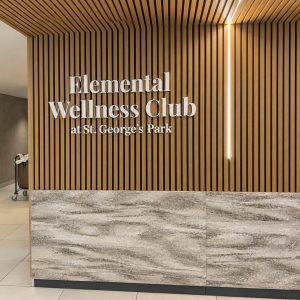 Reception area with a wood slat wall and illuminated sign reading 'Elemental Wellness Club at St. George's Park' above a marble front desk.