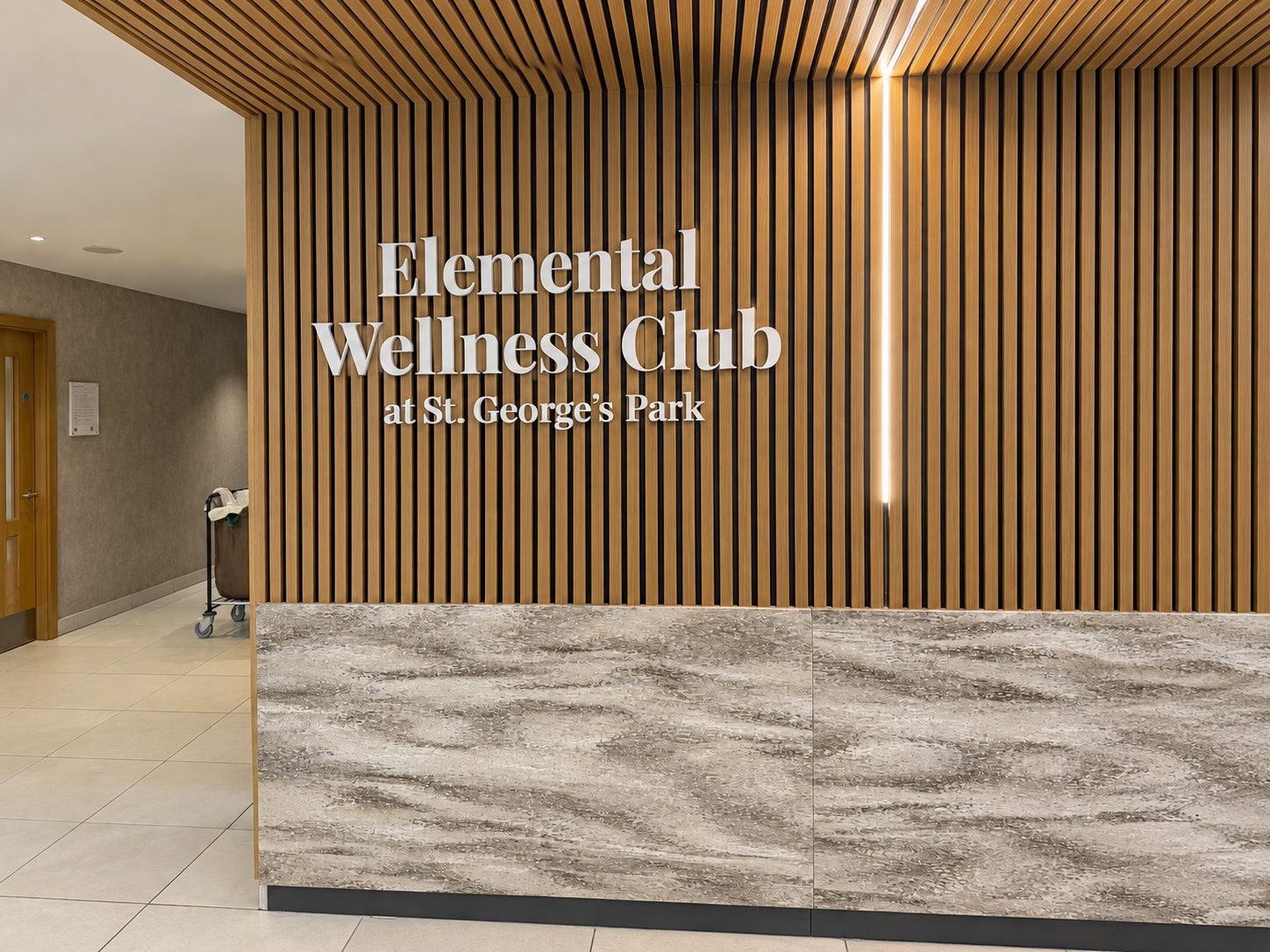 Reception area with a wood slat wall and illuminated sign reading 'Elemental Wellness Club at St. George's Park' above a marble front desk.