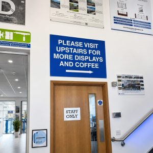 Blue sign reading 'PLEASE VISIT UPSTAIRS FOR MORE DISPLAYS AND COFFEE' with an arrow, mounted above a wooden 'Staff Only' door in a lobby area with an escalator to the right.