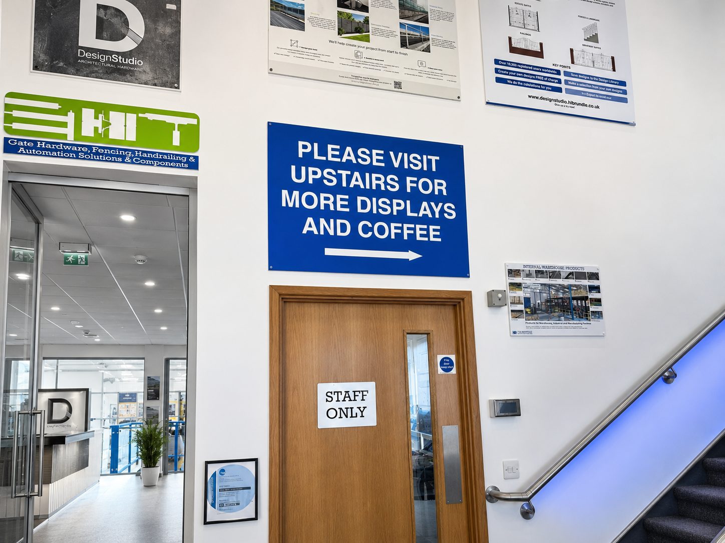 Blue sign reading 'PLEASE VISIT UPSTAIRS FOR MORE DISPLAYS AND COFFEE' with an arrow, mounted above a wooden 'Staff Only' door in a lobby area with an escalator to the right.