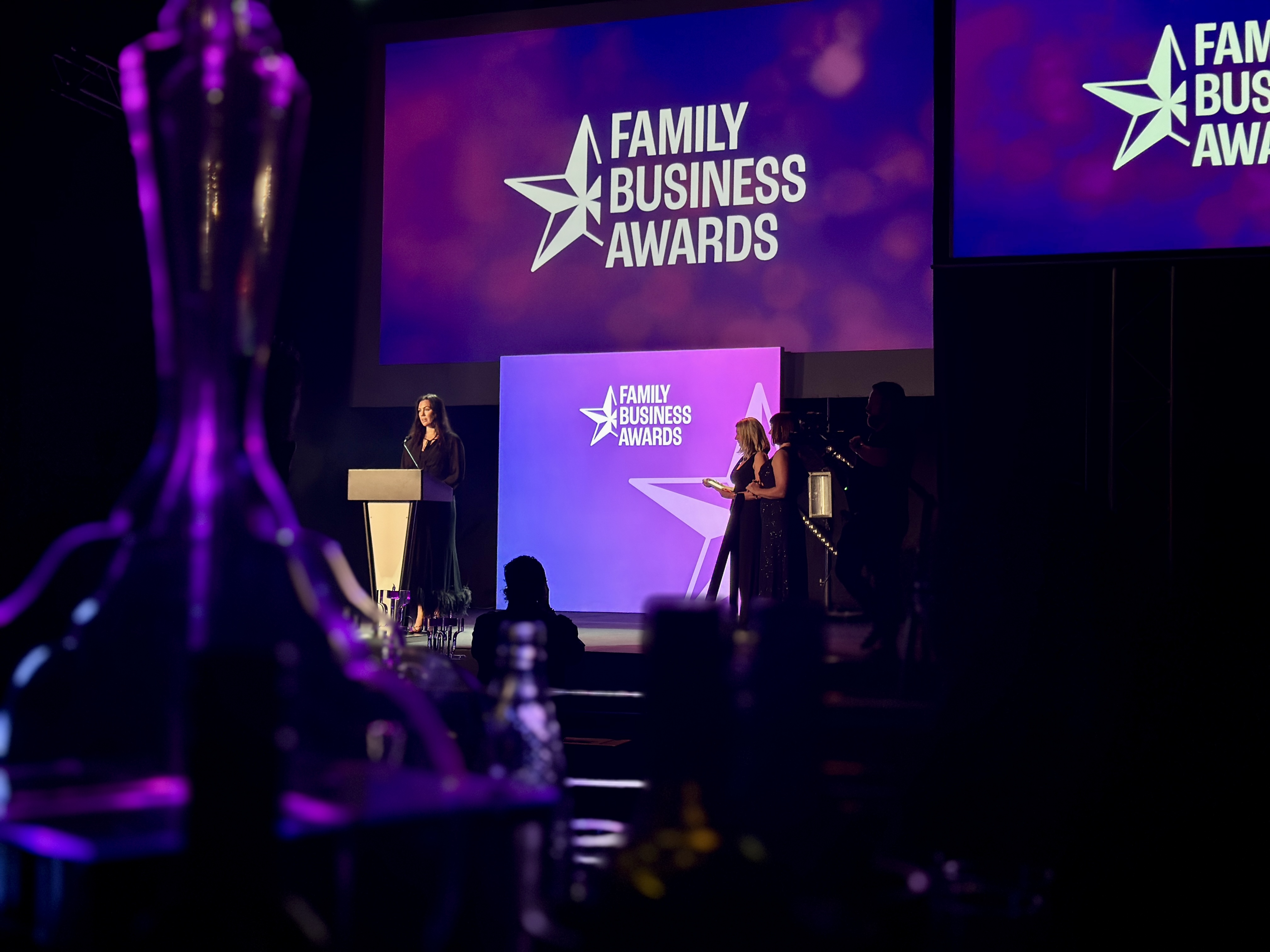 Stage at a Family Business Awards ceremony with a speaker at a podium and large purple screens displaying the event branding, a tall trophy in the foreground, and people on stage in the background.