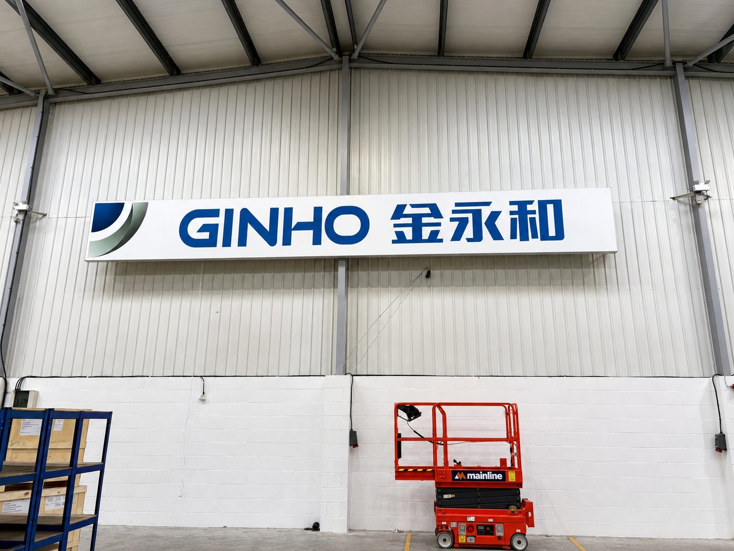 Industrial warehouse interior with a white sign reading GINHO and Chinese characters, and a red scissor lift nearby.