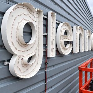 Close-up of a large white 3D illuminated sign mounted on a gray metal wall; wires and LED modules are visible behind the letters, with an orange scaffold in the foreground.