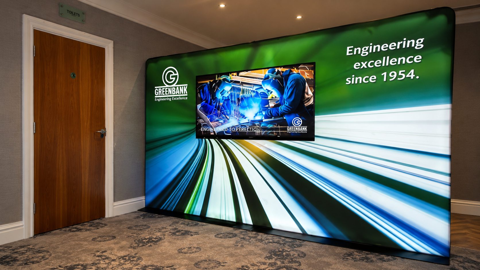 Large Greenbank Engineering display in a lobby, with a monitor showing welders and a door labeled toilets to the left.