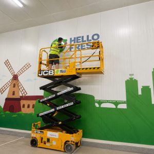 Worker on a yellow scissor lift applying the words HELLO FRESH to a wall with a windmill and green city skyline mural inside a warehouse, high-visibility vest on.