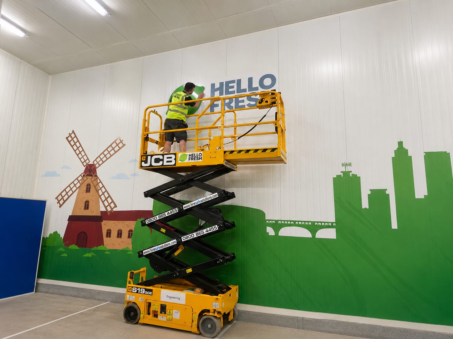 Worker on a yellow scissor lift applying the words HELLO FRESH to a wall with a windmill and green city skyline mural inside a warehouse, high-visibility vest on.