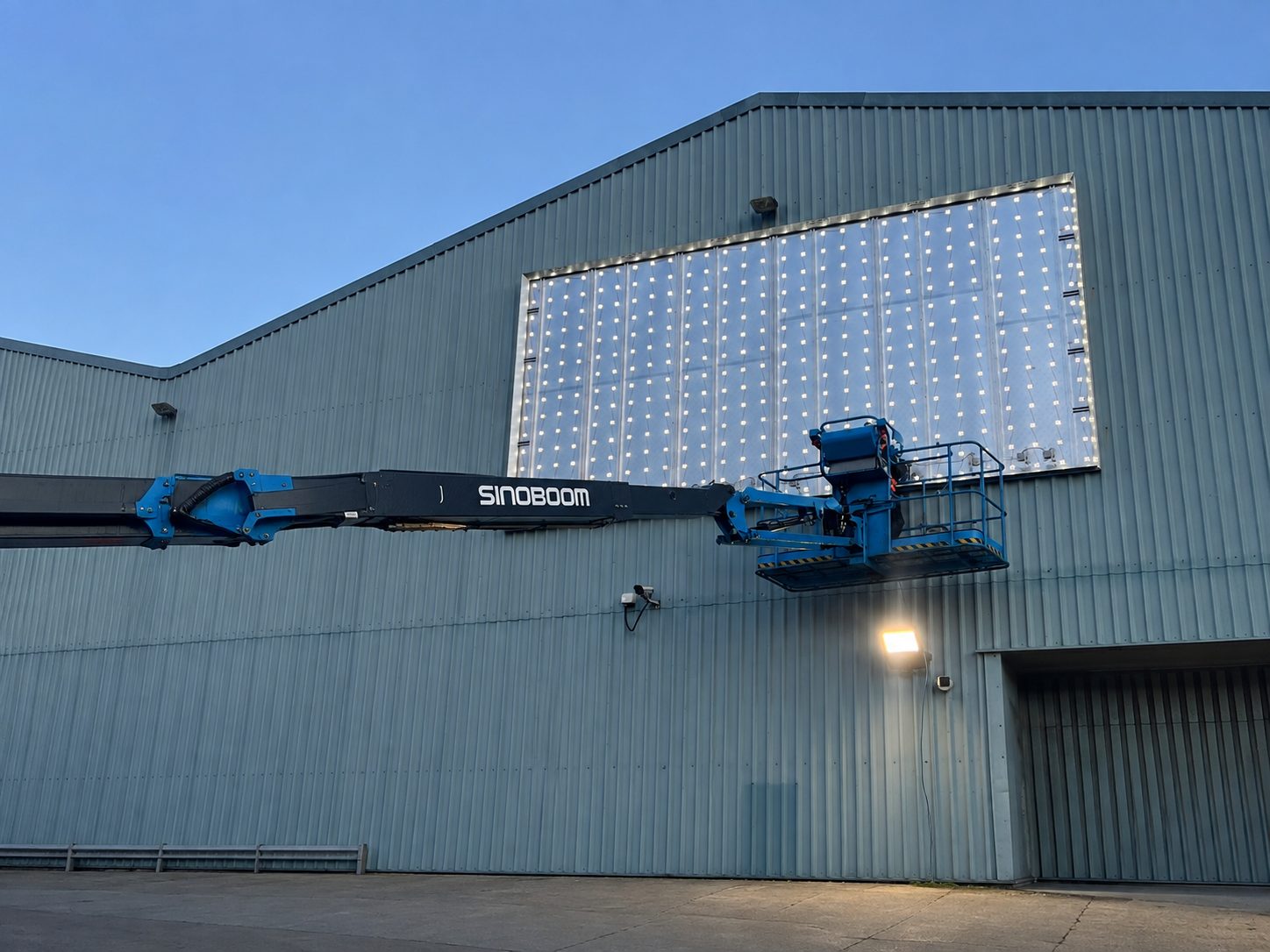 Blue Sinoboom hydraulic boom lift extends from the ground toward a large lit window on a gray metal warehouse exterior, suggesting maintenance work.