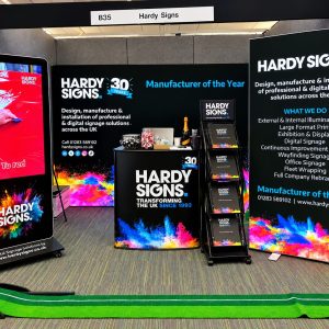 Trade show booth for Hardy Signs with black backdrop, colorful splash graphics, brochures, and a tall digital display on the left showing 'HARDY SIGNS'.