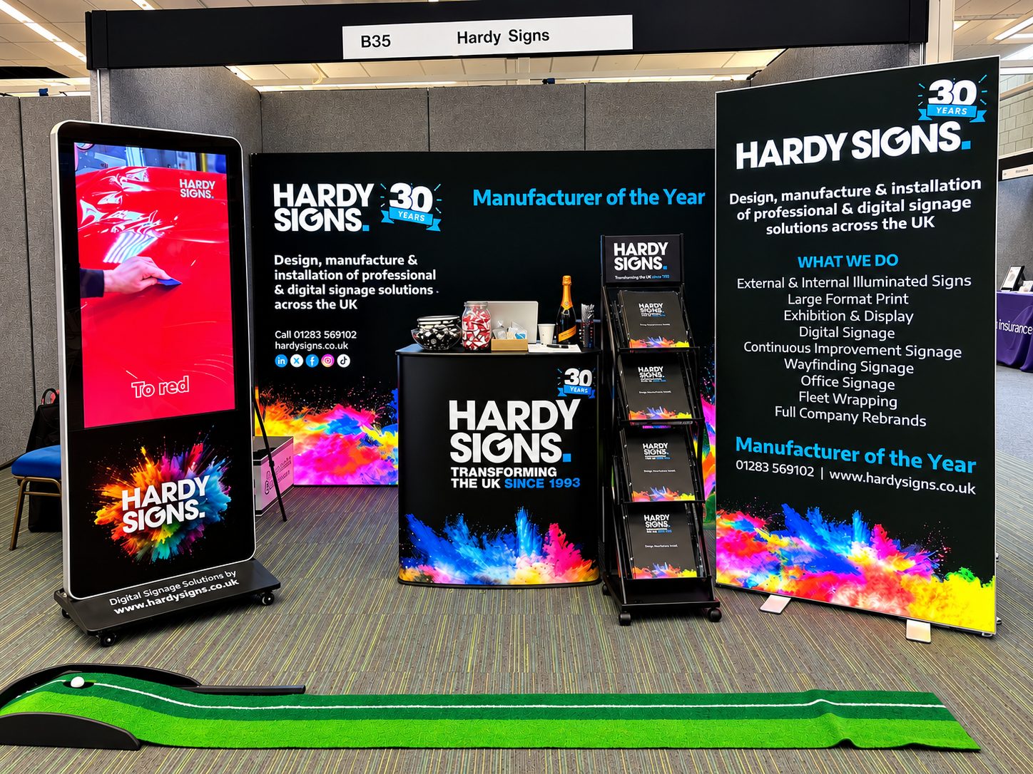Trade show booth for Hardy Signs with black backdrop, colorful splash graphics, brochures, and a tall digital display on the left showing 'HARDY SIGNS'.