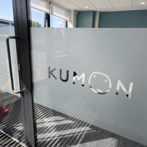Glass door with a frosted panel displaying the Kumon logo and a smiling 'O'; interior entry area with a metal handle visible and outdoor lot beyond.