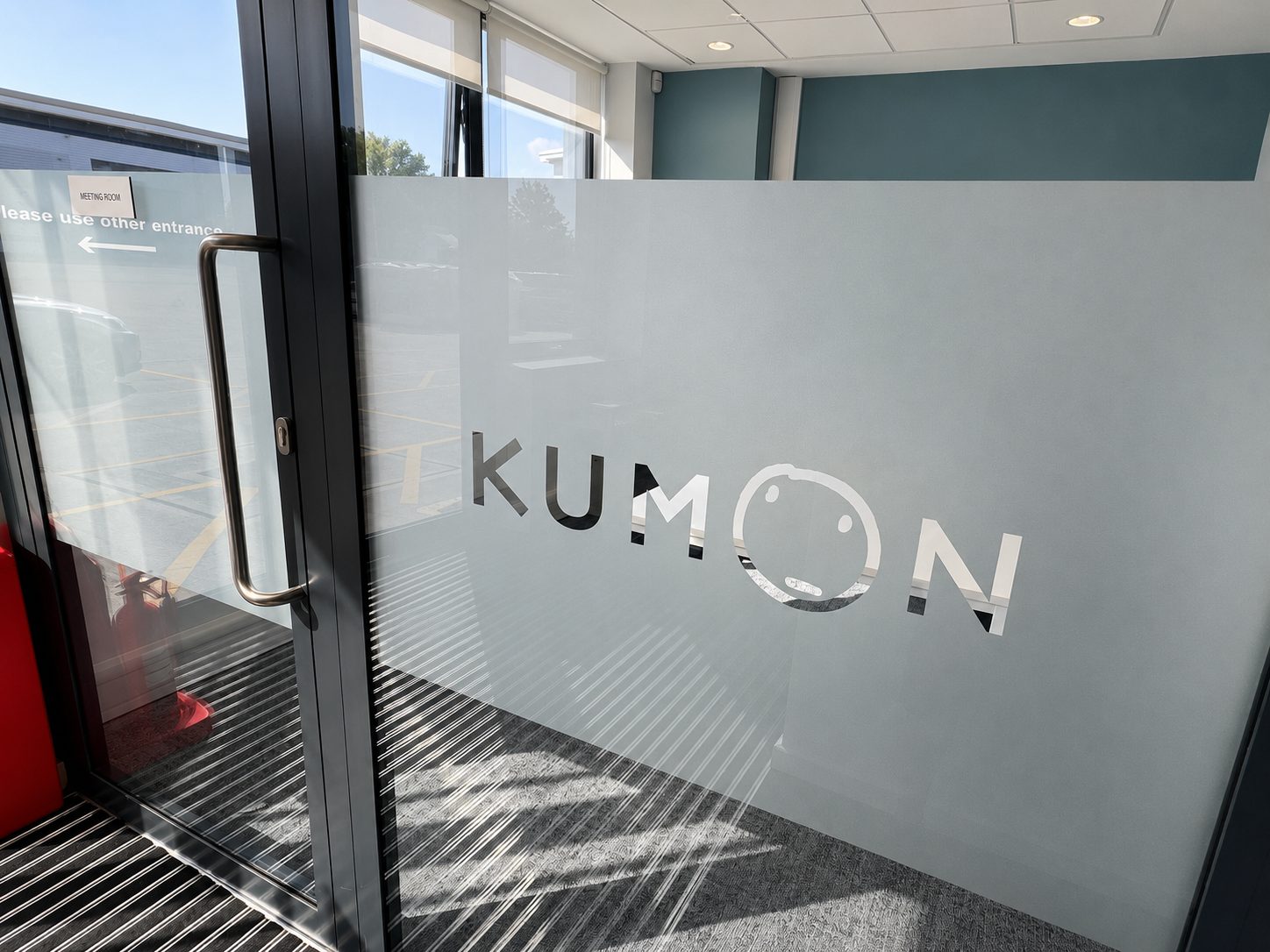 Glass door with a frosted panel displaying the Kumon logo and a smiling 'O'; interior entry area with a metal handle visible and outdoor lot beyond.