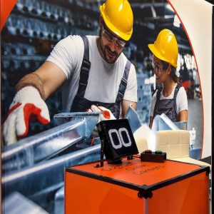 Trade show booth with workers in yellow hard hats and overalls on a large banner, an orange demonstration counter, and a digital display showing 00.