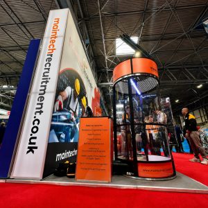 Trade show booth for Maintech Recruitment with a tall vertical sign reading 'maintech recruitment' and an orange information panel listing roles like maintenance and design engineers in a busy expo hall.