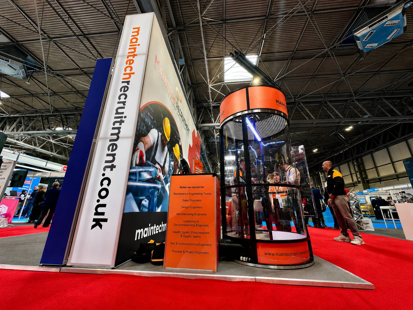 Trade show booth for Maintech Recruitment with a tall vertical sign reading 'maintech recruitment' and an orange information panel listing roles like maintenance and design engineers in a busy expo hall.