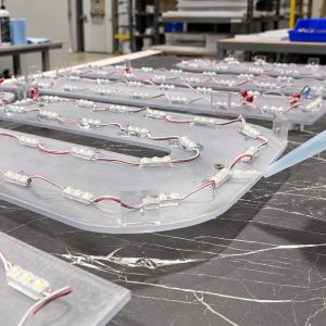 LED strip modules wired with red/white wires laid out on curved metal panels in a workshop; a gloved hand uses a caulk gun to apply adhesive.