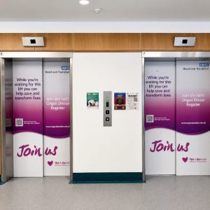 Hospital corridor with two elevator doors covered in NHS organ donation banners reading 'Join us' and 'Yes I donate'.