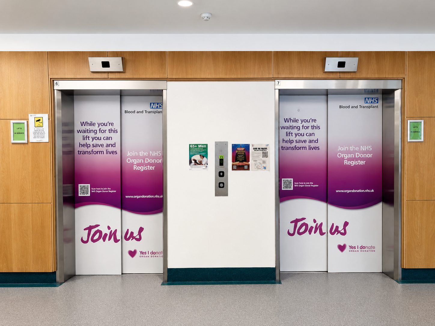 Hospital corridor with two elevator doors covered in NHS organ donation banners reading 'Join us' and 'Yes I donate'.