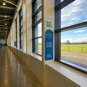 Industrial hallway with large windows looking onto a green field; safety poster on a pillar reads 'We care about safety' and 'Stop the machine before any intervention'.