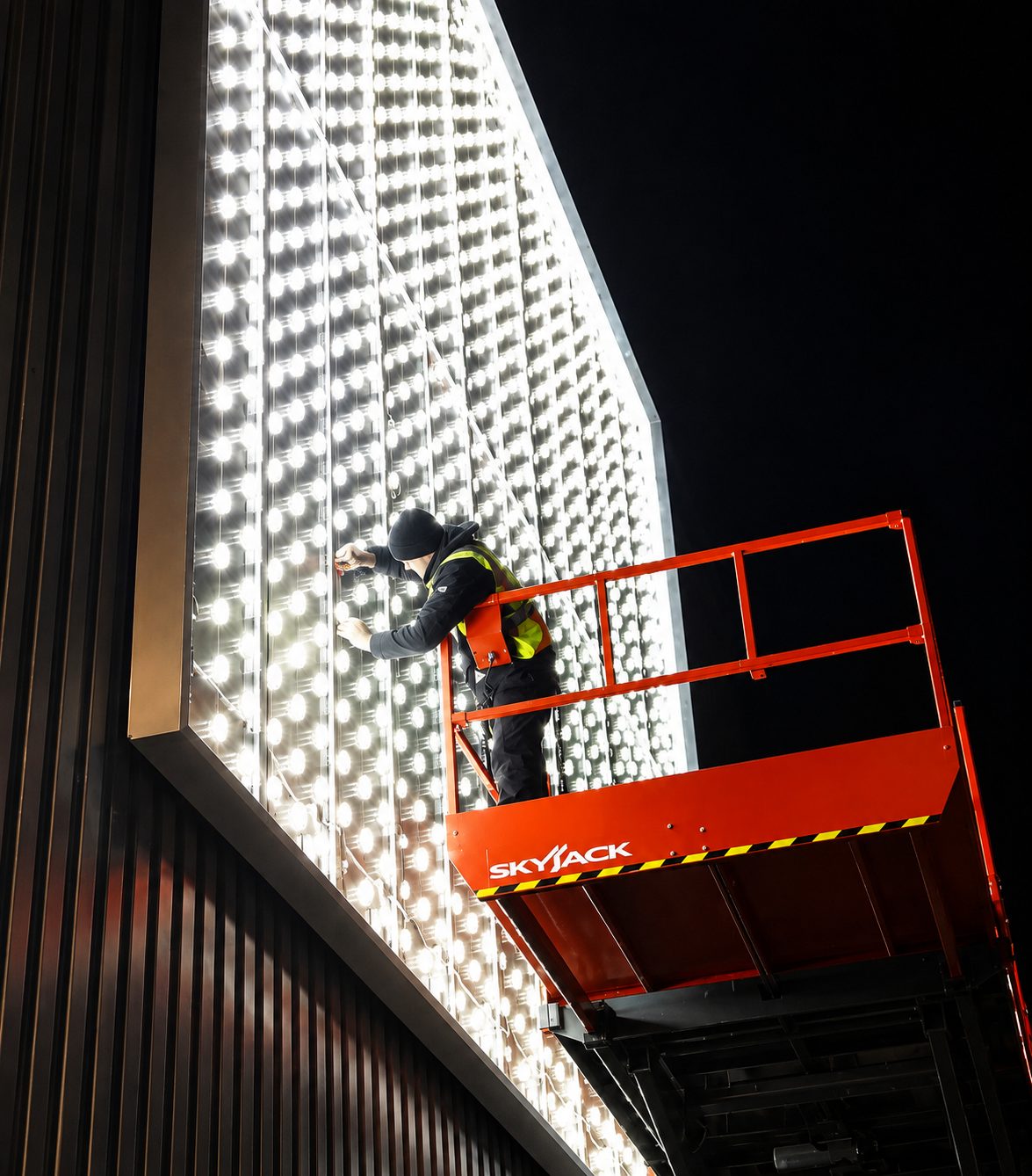 Worker on a red SkyJack lift adjusting a tall wall of bright LEDs at night.