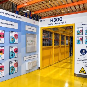 Safety check point entry with yellow framed gate labeled H300 in a warehouse. Posters and safety sheets on blue walls.
