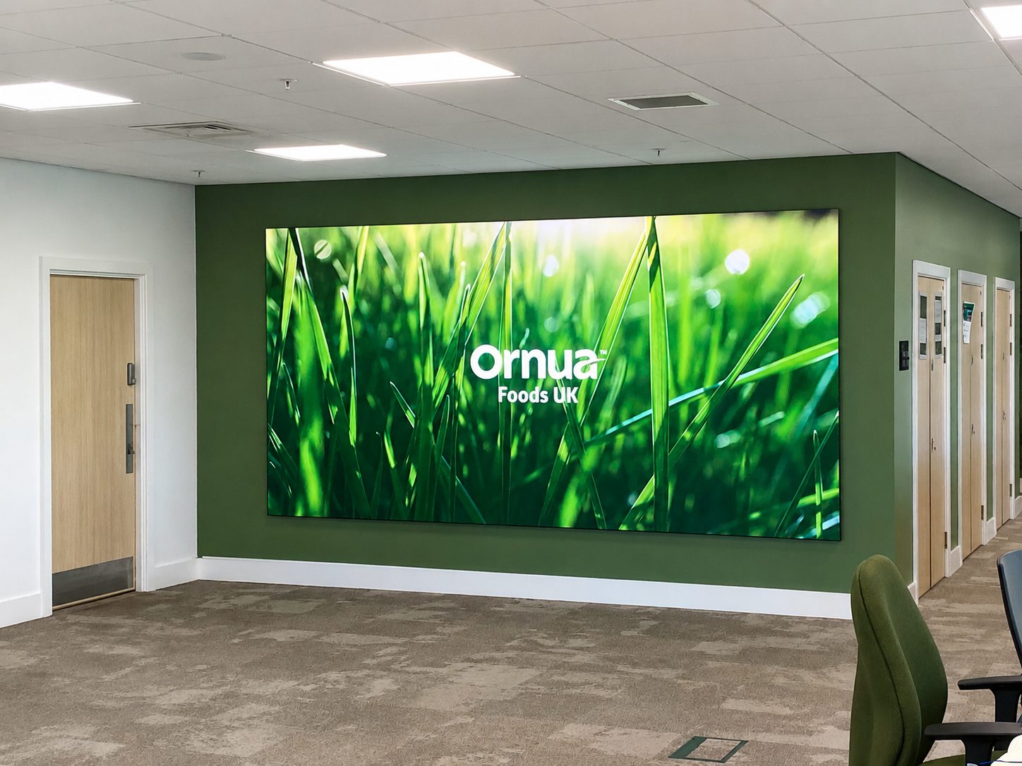 Large wall-mounted screen in a green-walled hallway displaying the Ornua Foods UK logo over a grass background, likely a lobby sign.