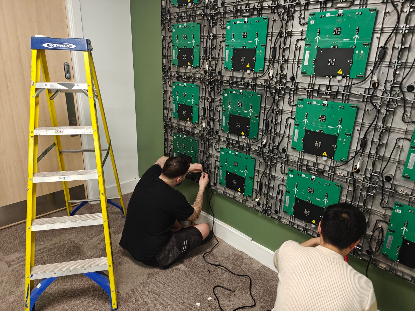 Two technicians kneel and work on a wall of green circuit-board panels connected by cables, with a yellow ladder nearby.