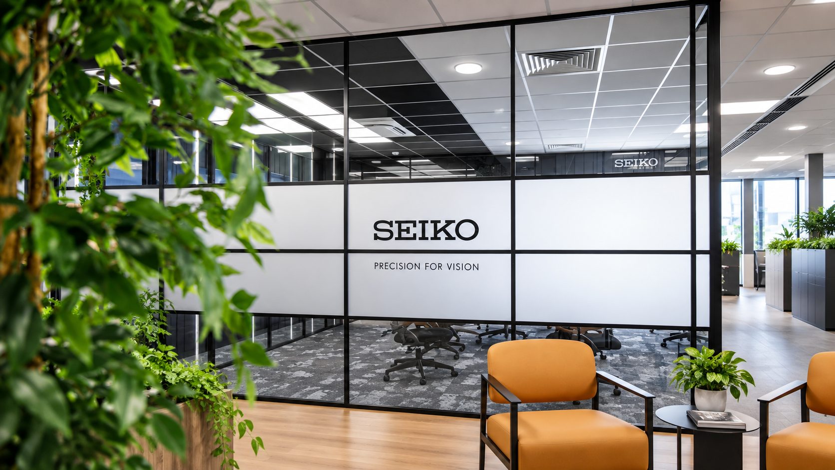 Office lobby with a glass partition displaying SEIKO branding and slogan, orange chairs, and surrounding green plants.