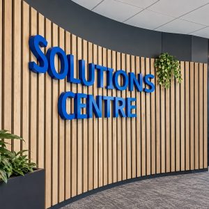 Curved wood-slat feature wall with bold blue 'SOLUTIONS CENTRE' lettering, plants, and glass-walled meeting room in a modern office entrance.