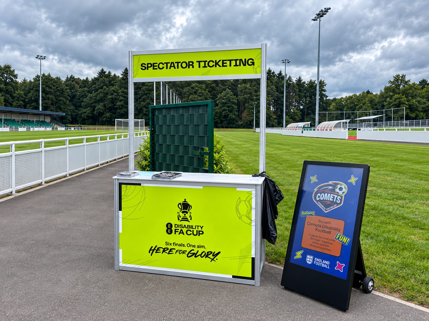 Bright yellow spectator ticketing booth at a football stadium with a bold SPECTATOR TICKETING sign and a large grassy field behind it.