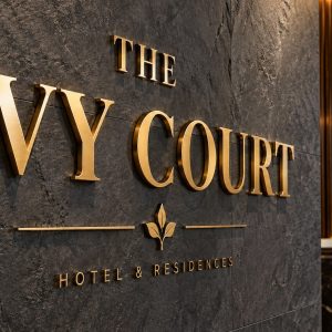 Gold letters spell 'THE IVY COURT' on a dark stone wall, indicating the hotel sign in a lobby. It reads 'Hotel & Residences' beneath a decorative emblem.
