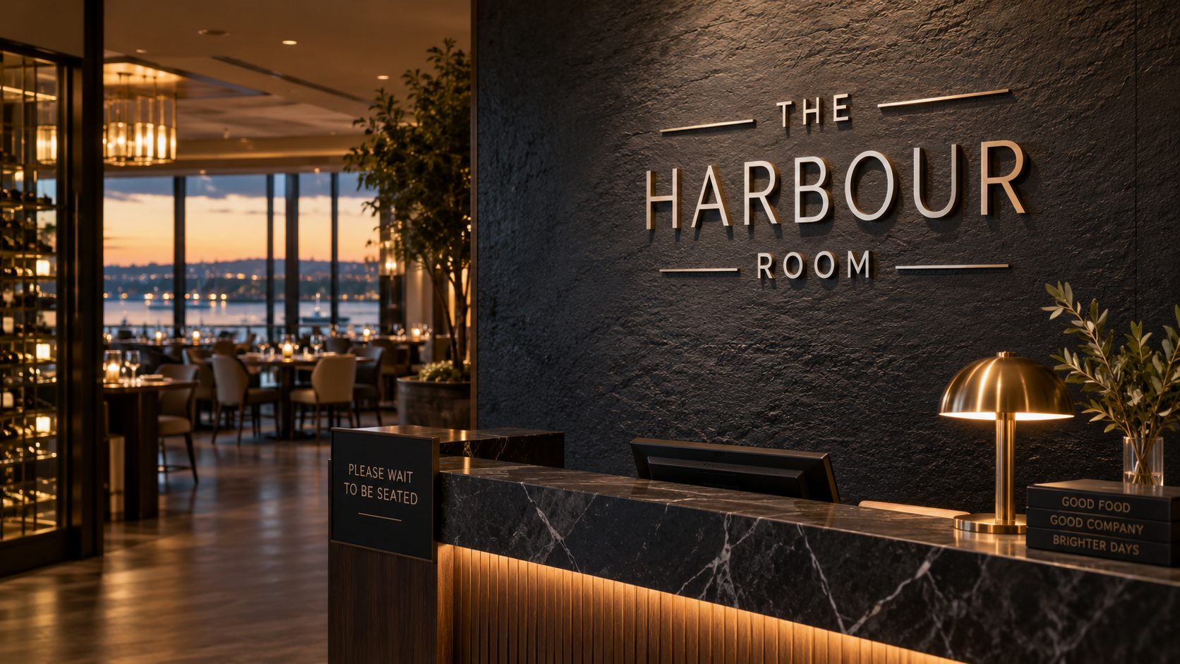 Reception area of The Harbour Room restaurant with a dark textured wall, illuminated sign, marble counter, and warm lighting by a harbor-view dining room