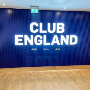 Blue wall with vertical blue slats and large white letters reading CLUB ENGLAND, with an illuminated exit sign overhead.
