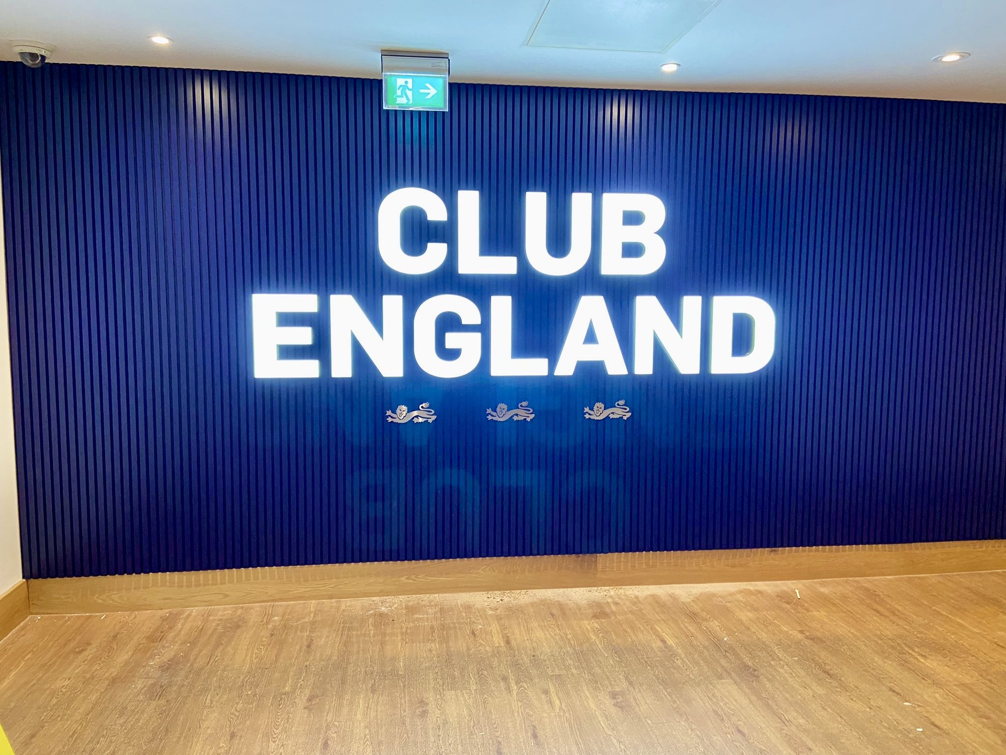 Blue wall with vertical blue slats and large white letters reading CLUB ENGLAND, with an illuminated exit sign overhead.