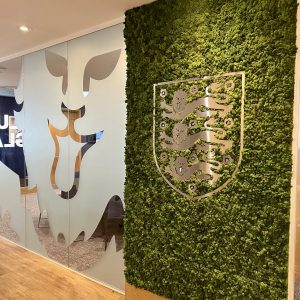 Office interior featuring a lush green moss wall with a silver crest emblem, next to frosted glass panels with leaf shapes and wooden flooring.