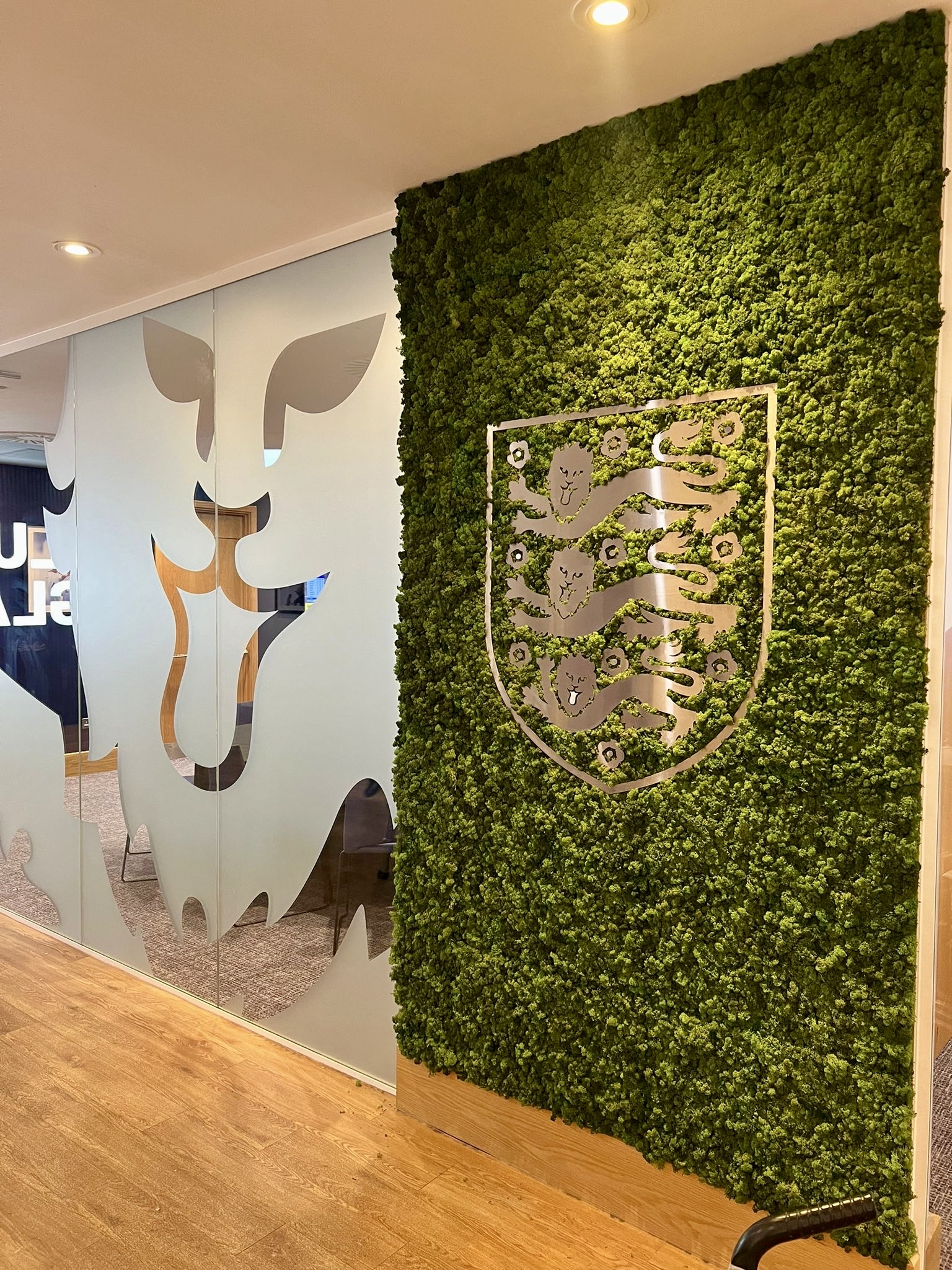 Office interior featuring a lush green moss wall with a silver crest emblem, next to frosted glass panels with leaf shapes and wooden flooring.