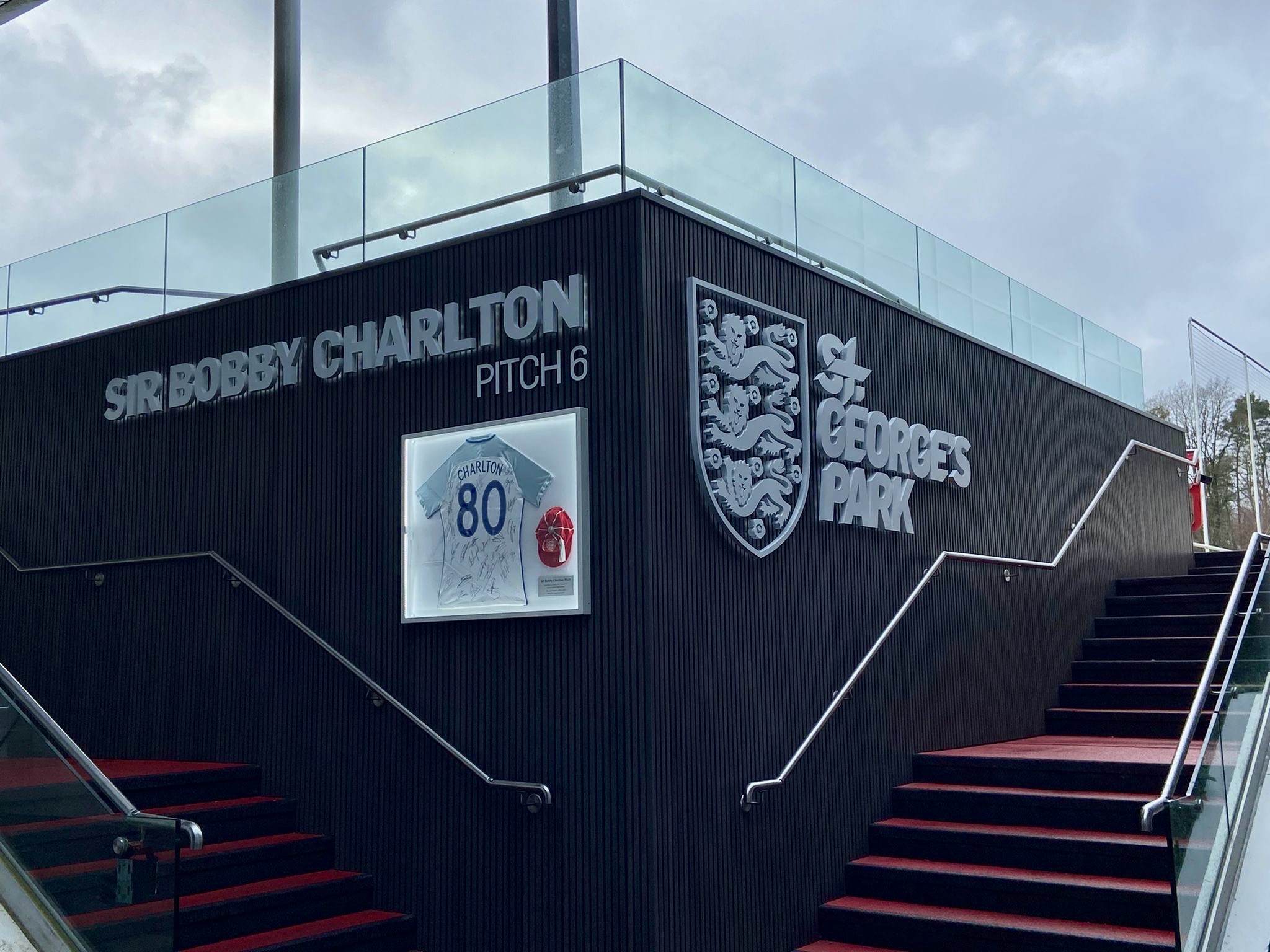Corner of a modern stadium building with a sign reading 'SIR BOBBY CHARLTON PITCH 6' and a visible crest, glass railing above, red-carpeted stairs below.
