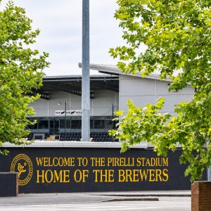 Sign reads: 'Welcome to the Pirelli Stadium, Home of the Brewers,' with green trees framing the entrance in the foreground.