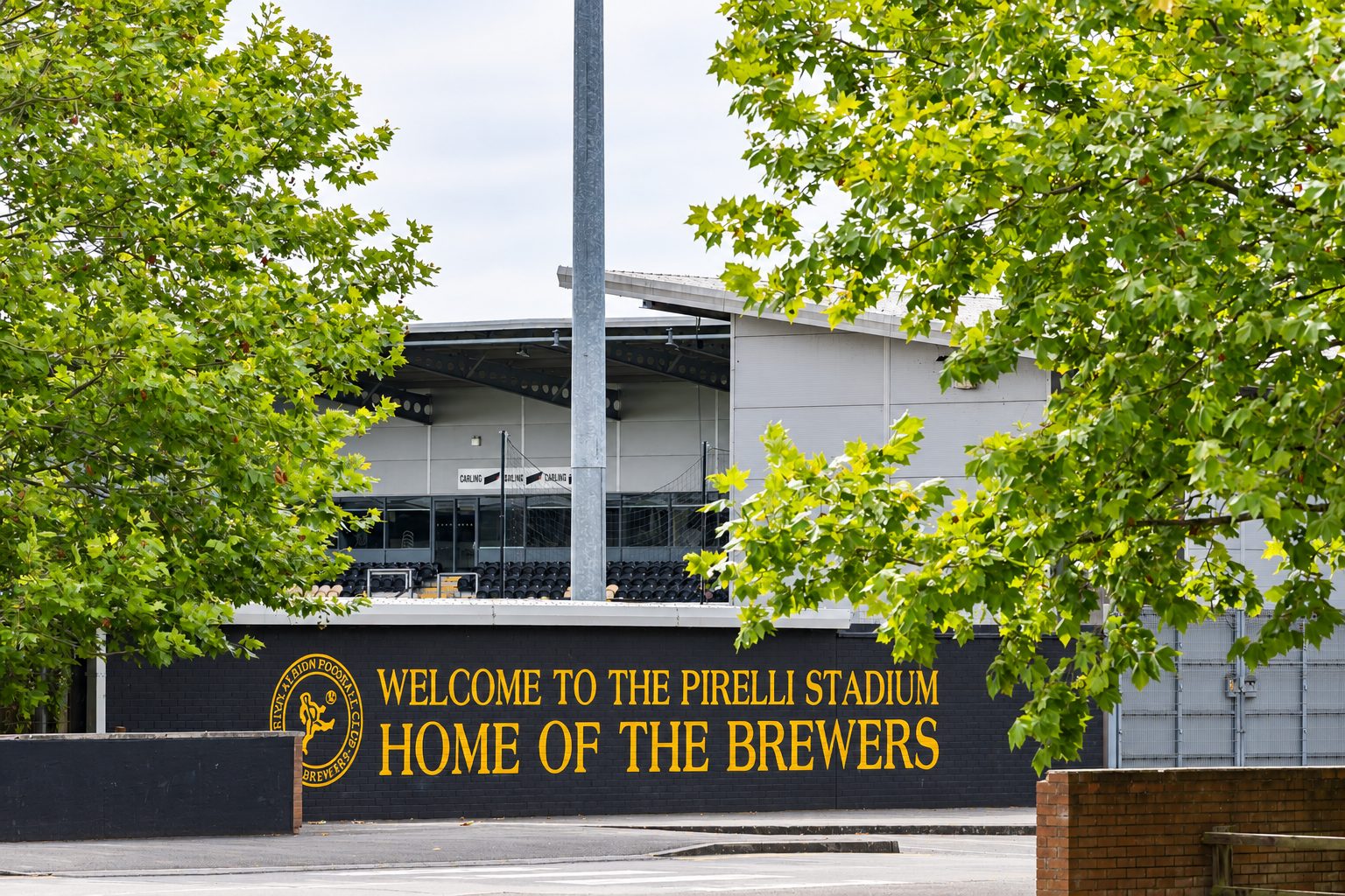 Sign reads: 'Welcome to the Pirelli Stadium, Home of the Brewers,' with green trees framing the entrance in the foreground.