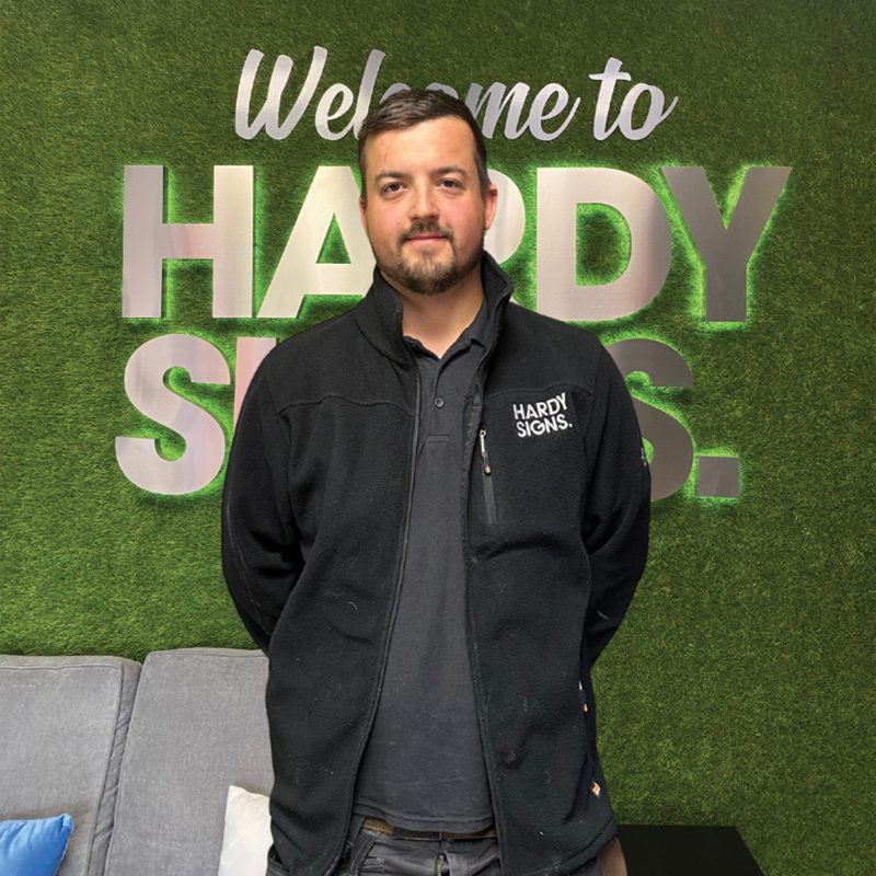 Man wearing a black Hardy Signs jacket stands in front of a green wall with large silver letters saying 'Welcome to HARDY SIGNS'.