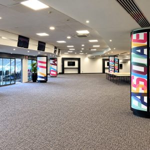 Expansive event hall with colorful cylindrical pillars displaying bold text, empty seating area, and a long row of tables in the back corner.—informational venue image
