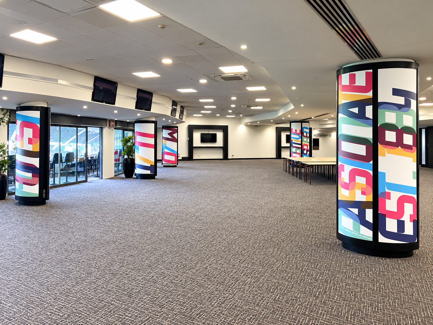 Expansive event hall with colorful cylindrical pillars displaying bold text, empty seating area, and a long row of tables in the back corner.—informational venue image
