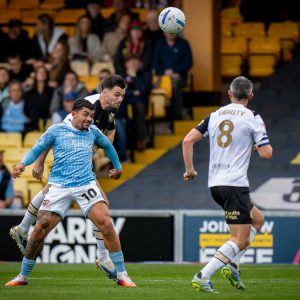 Soccer players chase a ball mid-air during a match in a stadium with yellow stands and crowd spectators far behind them as they compete for possession.