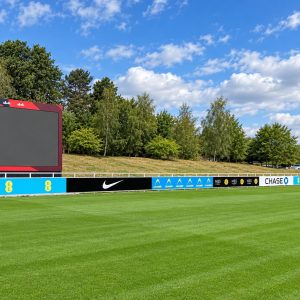 Wide view of a sports field with a large blank digital scoreboard and advertising boards along the sideline, blue sky above and trees in the background.
