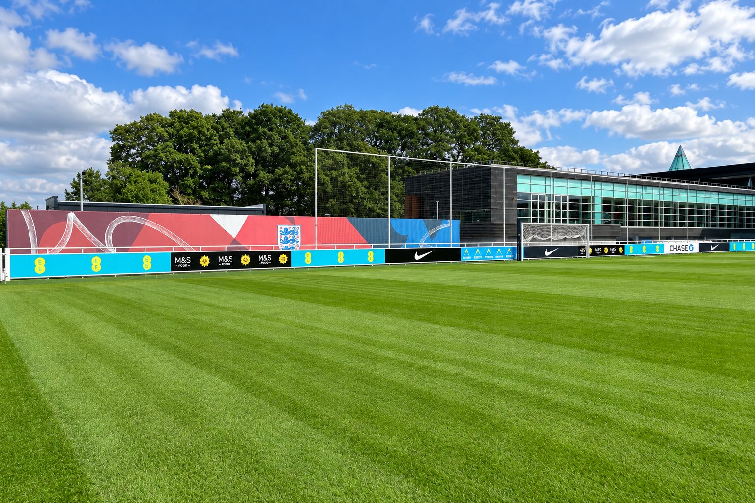 Soccer training field with green grass, advertising boards along a fence, and a modern glass-fronted building behind under a blue sky.