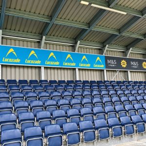 Rows of blue stadium seats under a metal roof with blue-and-yellow Lucozade banners and M&S Food signage along the front.