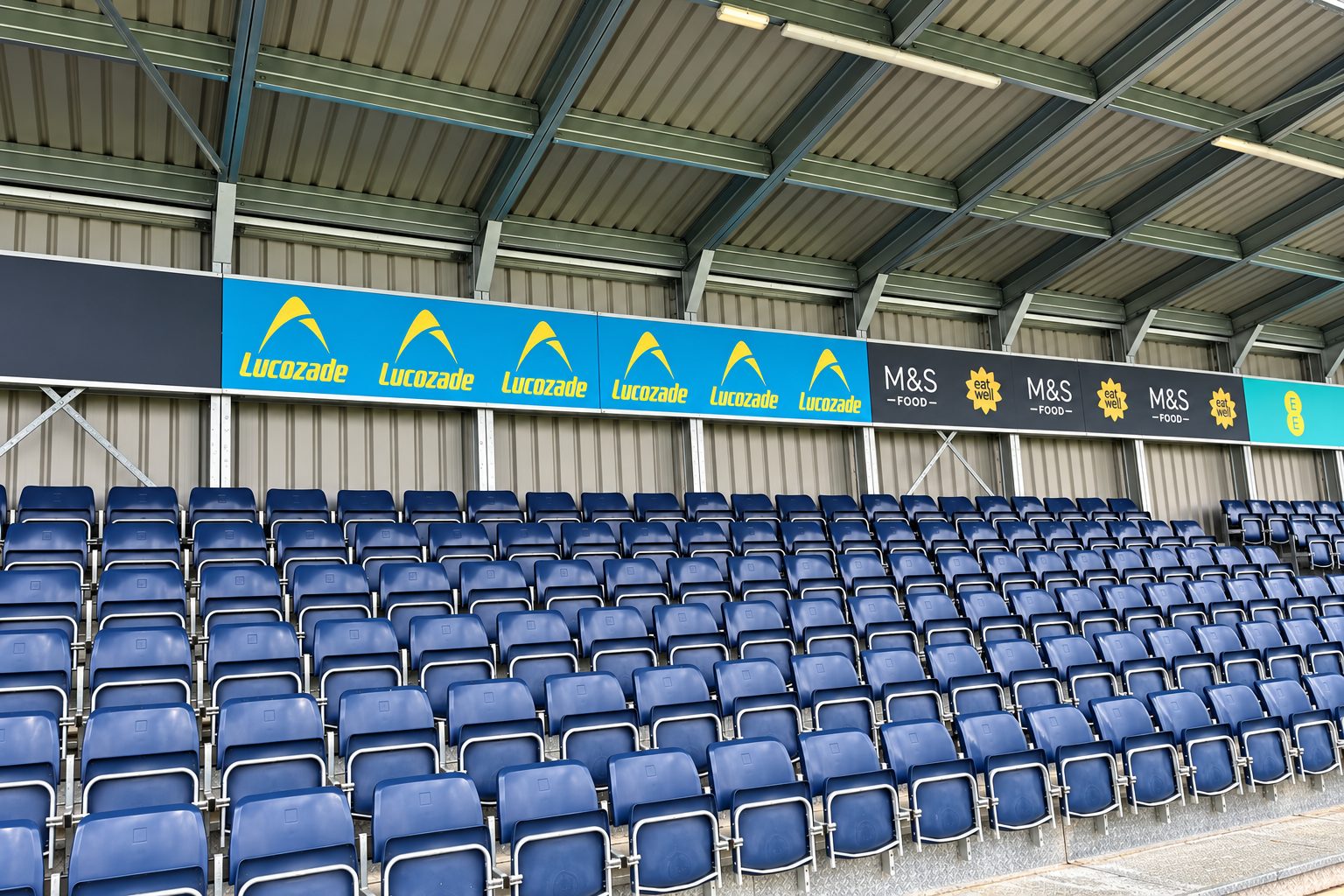 Rows of blue stadium seats under a metal roof with blue-and-yellow Lucozade banners and M&S Food signage along the front.