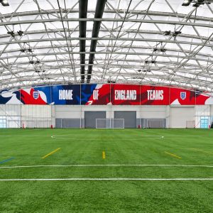 Indoor artificial turf soccer field with a large banner reading 'HOME OF ENGLAND TEAMS' along the back wall.