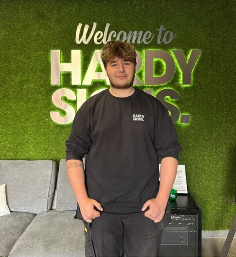 Young man in a black long-sleeve shirt standing in front of a green wall with 'Welcome to HARDY SIGNS' text, posing for a staff photo.