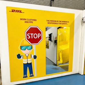 Bright yellow safety panel with a cartoon worker holding a Stop sign and wearing PPE; DHL logo at top left, warning text about work clothing and PPE